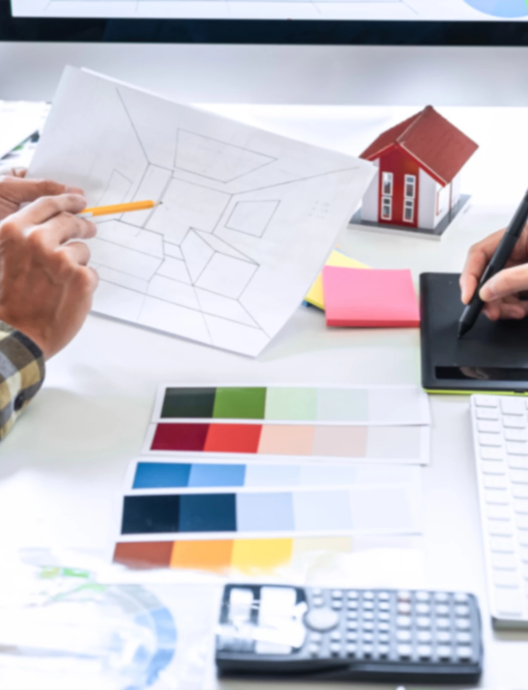 m�o segurando um l�pis apontando para desenho, paleta de cores em cima da mesa, maquete de casa, calculadora, mesa digitalizadora e papeis adesivos